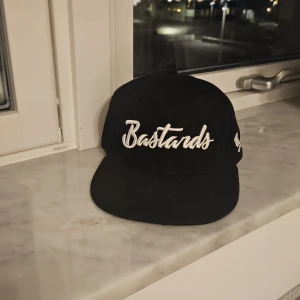 Svart keps Bastard Burgers - Svart snapback-keps från Bastard Burgers med vit broderad text 'Bastards' framtill. Kepsen har platt skärm och justerbar baksida. Insidan har coola små vita ikoner längs sömmarna. Perfekt för dig som gillar streetwear och vill sticka ut.
