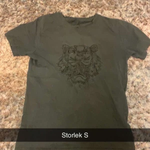 Kenzo T-shirt - Snygg mörkgrön t-shirt i storlek S med ett stort tigertryck på bröstet. T-shirten har rund hals och korta ärmar, perfekt för dig som gillar djurmotiv och vill sticka ut lite extra. Passar dig som gillar streetstyle och coola detaljer.