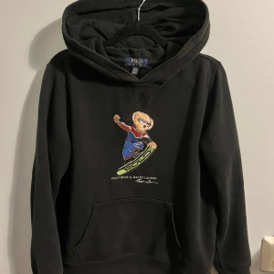 Svart hoodie Polo Ralph Lauren björn - Svart hoodie från Polo Ralph Lauren med ikonisk Polo Bear-tryck på bröstet. Tröjan har huva, känguruficka och långa ärmar. Perfekt för dig som gillar streetstyle och vill ha något bekvämt och snyggt.