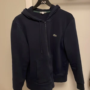 Marinblå hoodie från Lacoste - Snygg marinblå hoodie från Lacoste med klassisk krokodillogo på bröstet. Tröjan har huva med snörning, dragkedja hela vägen och två stora fickor framtill. Mjuk insida och ribbade muddar vid ärmslut och nederkant. Perfekt för chill dagar.