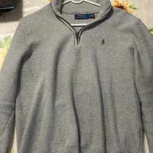 Grå stickad tröja Polo Ralph Lauren - Snygg grå stickad tröja från Polo Ralph Lauren med half zip och klassisk krage. Tröjan har långärmad passform och diskret logga broderad på bröstet. Perfekt för dig som gillar stilrena och tidlösa plagg med premiumkänsla.