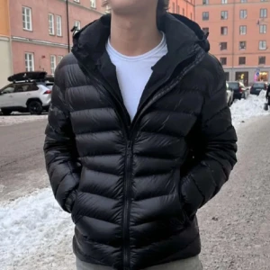 Svart Colmar pufferjacka med huva - Säljer en svart pufferjacka från Colmar med glansig finish och huva. Jackan har horisontella quiltade paneler, dragkedja framtill och två sidofickor. Märkeslogga på vänster ärm. Perfekt för kalla dagar och har en riktigt clean look. Originalpris 6500kr