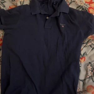 Marinblå pikétröja från GANT - Snygg marinblå pikétröja från GANT med klassisk krage och två knappar framtill. Diskret logga broderad på bröstet. Perfekt för dig som gillar stilrena och tidlösa plagg. Skön bomullskvalitet och korta ärmar.