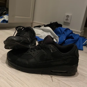 Svarta Nike Air Max 90 sneakers - Säljer ett par helsvarta Nike Air Max 90 sneakers med klassisk chunky sula och synlig Air-enhet. Skorna har mesh och skinnpaneler, rund tå och snörning. Perfekt för dig som gillar streetwear och vill ha en stilren look.