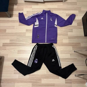 Svarta Adidas Real Madrid träningsbyxor - Svarta träningsbyxor från Adidas med Real Madrid-logga på benet och klassiska vita ränder längs sidorna. Byxorna har elastisk midja och smal passform, perfekta för sport eller chill. Materialet är troligtvis polyester och designen är modern och sportig. Storleken är 164/176 i barn storlek!!! Inte män. Den kanske också är lite mindre för att jag tvätta den.