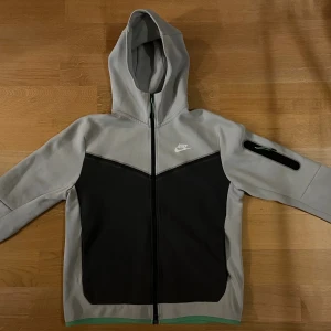 Nike grå och svart zip hoodie M - Nike zip hoodie i grått och svart med huva och hel dragkedja framtill. Hoodie har en cool design med svart V-formad panel och en ficka med dragkedja på ärmen. Perfekt för dig som gillar sportig stil och vill ha något som sticker ut. Priset kan diskuteras vid snabb affär 💯 