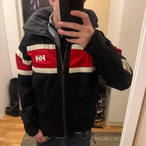 Helly Hansen fodrad jacka  - Säljer en Helly Hansen jacka i mörkblå med röda och vita ränder över bröst och armar. Jackan har huva, dragkedja framtill och fleecefoder i kragen. Regular fit och snygga HH-loggor på bröst och ärm.