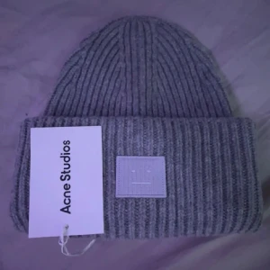 Acne mössa - Säljer en grå ribbstickad mössa från Acne Studios med det ikoniska face-patch framtill. Mössan är i mjuk ull och har en bred uppvikt kant. Perfekt accessoar för kalla dagar och snygg till streetwear eller casual outfits.