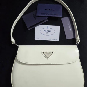 Vit axelväska från Prada i skinn - Snygg vit axelväska från Prada med klassisk, stilren design och silverfärgad logotyp på framsidan. Väskan är tillverkad i slätt skinn och har en enkel axelrem. Perfekt för dig som gillar minimalistisk och lyxig stil.Den har några fläckar på baksidan, som syns på bilderna. Jag köpte den från Vinted.