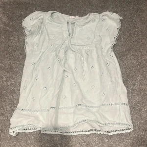 Ljusblå blus med broderade detaljer från GAP - Supersöt ljusblå blus från GAP med broderade hålmönster och volangärmar. Blusen har knytband fram vid halsen och fina spetsdetaljer längs kanterna. Luftig och skön i bomull, perfekt för varma dagar. Snygg till jeans eller kjol!