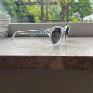 Di blåe transparenta solglasögon - Snygga solglasögon från Di blåe med transparent båge och runda, mörkgrå linser. Modern och clean design som ger en cool vibe. Bågarna är gjorda i syntetmaterial och har en diskret blå logga på sidan. Perfekt för soliga dagar utomhus.
