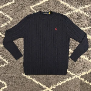 Polo Ralph Lauren tröja Marinblå - Snygg marinblå stickad tröja från Polo Ralph Lauren med klassiskt kabelstickat mönster och röd broderad logga på bröstet. Rund halsringning och långa ärmar. Jag är 180cm, 68kg och den passar mig perfekt.