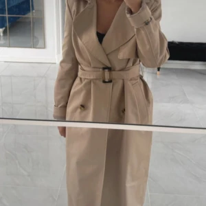 Beige trenchcoat med bälte - Snygg beige trenchcoat med klassisk krage och dubbelknäppning. Jackan har ett matchande bälte i midjan och långa ärmar med justerbara spännen vid handlederna. Perfekt för dig som gillar stilrena och tidlösa plagg.