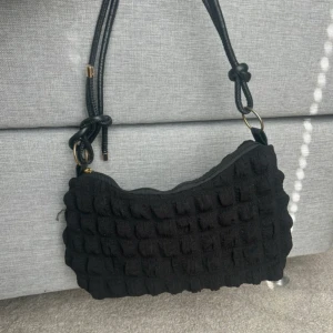 Svart quiltad axelväska med dragkedja - Snygg svart axelväska med quiltad struktur och justerbar axelrem i repdesign. Väskan har en praktisk dragkedja i guld och ett ljust innerfoder. Perfekt storlek för det viktigaste och ger en cool touch till din outfit.