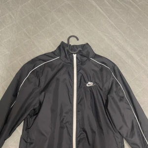 Svart vindjacka från Nike - Svart vindjacka från Nike med vit dragkedja och vita detaljer längs axlarna. Jackan har hög krage och klassisk Nike-logga på bröstet. Tillverkad i lätt polyester som är perfekt för blåsiga dagar. Snygg och sportig look med lång ärm.