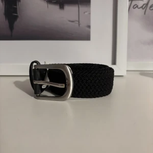 Svart flätat bälte med silverfärgat spänne - Snyggt svart flätat bälte med ett stilrent silverfärgat metallspänne. Bältet är tillverkat i polyester och har en klassisk, enkel design som passar till jeans eller chinos. Perfekt längd på 70 cm och smidig att justera efter behov.