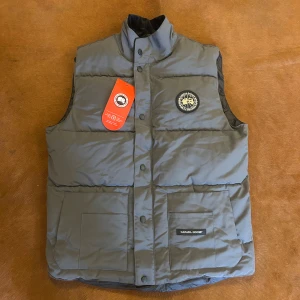 Grå dunväst från Canada Goose - Snygg grå dunväst från Canada Goose med hög krage och tryckknappar framtill. Västen har två stora fickor med logotyp på ena fickan och ett broderat märke på bröstet. Perfekt för kalla dagar och har en stilren, quiltad design.