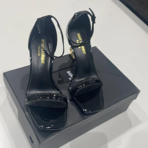 Svarta Saint Laurent sandaler med YSL-klack - Svarta sandaler från Saint Laurent med glansig finish och fyrkantig tå. Skorna har en öppen tå, smal rem över foten med glittrig detalj och justerbar ankelrem. Klacken är i guld med YSL-logga, vilket ger en lyxig vibe. Perfekta för dig som gillar statement heels.