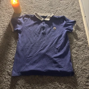 Blå Moncler pikéskjorta med vit krage - Blå pikéskjorta från Moncler med vit krage och vita muddar vid ärmslut. Skjortan har korta ärmar, knappar framtill och Moncler-logga broderad på bröstet. Materialet är mjuk bomull som känns skönt mot huden. Perfekt för dig som gillar stilrena märkesplagg.