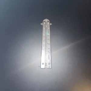 En utvecklingsbar kam  - Bilderna visar en butterfly-kam i metall, designad som en balisong. Produkten är inte elektronik och har inga synliga elektroniska funktioner, specifikationer eller märke. Kammen verkar vara i gott skick med mindre slitage, och är ett roligt och praktiskt tillbehör för styling.