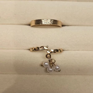 Två guldiga ringar med pärldetalj - Säljer två snygga guldiga ringar. Den ena har ett diskret mönster och den andra är tvinnad med tre små vita pärlor som hänger som en charm. Perfekt för dig som gillar unika accessoarer med lite extra detalj.