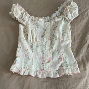 Blommig blus med volang och puffärm - Supersöt blommig blus i ljusa färger med rosa och gröna detaljer. Toppen har puffiga korta ärmar, volangkant nertill och knappar framtill. Smockad rygg för skön passform och romantisk vibe. Perfekt för dig som gillar en feminin och trendig stil.