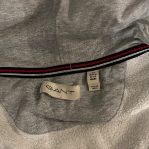 Grå hoodie från GANT med dragkedja - Snygg grå hoodie från GANT med huva och hel dragkedja framtill. Tröjan har två fickor och ett litet broderat GANT-emblem på bröstet. Insidan har en randig detalj i rött, blått och vitt. Perfekt för chill dagar och enkel att matcha. 