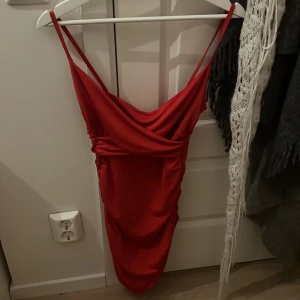 Röd omlottklänning med drapering - Säljer en röd kortklänning med smala axelband och snygg omlott-drapering framtill. Klänningen har en tight passform och rynkade detaljer längs ryggen för en extra snygg siluett. Perfekt för dig som vill sticka ut med en färgstark look.