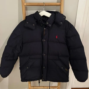 Mörkblå pufferjacka från Polo Ralph Lauren - Säljer en mörkblå pufferjacka från Polo Ralph Lauren med huva, dragkedja och knappar framtill. Jackan har två stora fickor med knappstängning och den klassiska röda Polo-loggan på bröstet. Perfekt för kalla dagar och riktigt snygg stil.