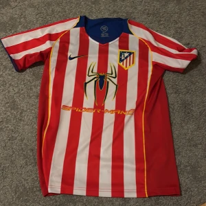 Atletico Madrid Spider-Man 2 fotbollströja - Säljer en Atletico Madrid fotbollströja från Nike med röda och vita ränder, blå detaljer och gula inslag. Framsidan har Spider-Man 2-tryck och klubbmärke, baksidan har F. Torres och nummer 9 i gult. Kortärmad och tillverkad i polyester.