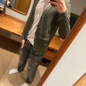 Olivgrön kofta från Polo Ralph Lauren - Olivgrön stickad kofta från Polo Ralph Lauren med snygg struktur, v-ringning och knäppning framtill. Två fickor framtill och ribbade muddar. Perfekt att slänga över en t-shirt för en chill look.