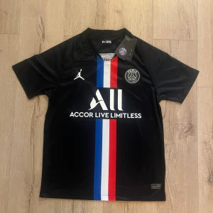 PSG x Jordan svart fotbollströja S - Svart PSG fotbollströja i samarbete med Jordan, tillverkad i lätt och ventilerande polyester. Framsidan har en vertikal rand i blått, vitt och rött, samt tryck med klubbmärke och Jordan-logga. Snygg V-ringning och korta ärmar, perfekt för dig som gillar streetwear och fotboll.