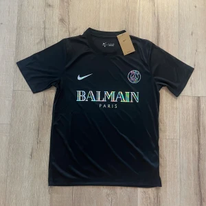 Svart PSG x Balmain fotbollströja Nike - Svart fotbollströja från Nike med Paris Saint-Germain x Balmain samarbete. Tröjan har korta ärmar, rund hals och är tillverkad i Dri-Fit material som andas. Framsidan har holografiskt Balmain-tryck och PSG-logga. Perfekt för dig som gillar exklusiva samarbeten.
