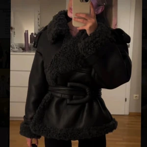 Adoore jacka - Säljer en svart teddyjacka från NA-KD med bred krage, fluffiga detaljer och snyggt bälte i midjan. Jackan har lång ärm, markerade axlar och coola knappar vid ärmslut. Perfekt för dig som vill ha en statement jacka med edge.