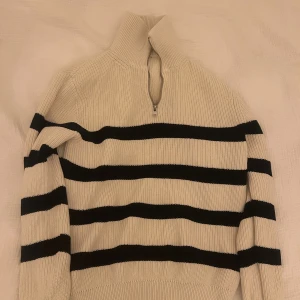 Randig stickad half zip tröja - Snygg stickad tröja med svartvita ränder och half zip-dragkedja i halsen. Tröjan har hög krage och långa ärmar, perfekt för dig som gillar en chill och stilren look. Passar till jeans eller cargo pants för en casual vibe.