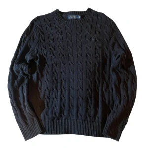 Mörkblå kabelstickad tröja Polo Ralph Lauren | M - Kabelstickad mörkblå tröja från Polo Ralph Lauren med klassisk rund halsringning och ribbade muddar. Tröjan har diskret broderad logga på bröstet och är tillverkad i mjuk bomull. Perfekt för dig som gillar en tidlös och clean stil.