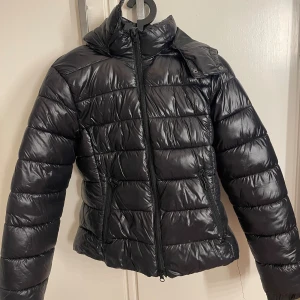 Svart pufferjacka med huva - Säljer en svart pufferjacka med glansig finish och huva. Jackan har quiltad design, två sidofickor med dragkedja och hel dragkedja framtill. Perfekt för kalla dagar och har en modern, lite boxig passform som är både snygg och bekväm.
