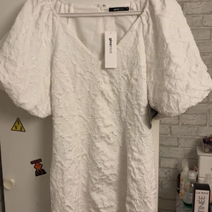 Vit brokadklänning Gina Tricot 36 - Supersnygg vit brokadklänning från Gina Tricot i storlek 36. Klänningen har puffiga korta ärmar, v-ringad hals och ett strukturerat tyg med brokadmönster. Dragkedja bak för enkel påklädning. Perfekt för dig som vill sticka ut med en elegant look.