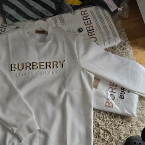 Vit Burberry sweatshirt med logga - Snygg vit sweatshirt från Burberry med klassisk logga och texten 'London England' på bröstet. Loggan har Burberrys ikoniska rutiga mönster i bokstäverna. Tröjan har rund hals, långa ärmar och ribbade muddar. Perfekt för dig som gillar stilrena designer och premiumkänsla.