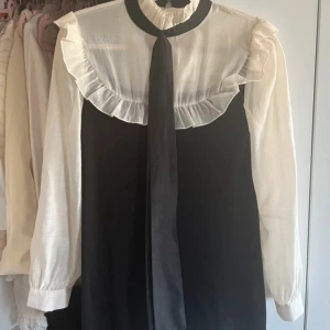 Svartvit klänning med volang och rosett - Säljer en svartvit klänning med långärmade vita ärmar och volangdetaljer vid axlar och hals. Klänningen har en svart rosett framtill och kontrasterande svart kjoldel. Perfekt för dig som gillar en chic och klassisk look.