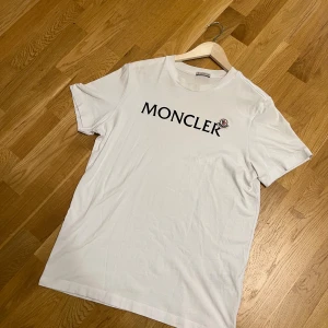 Vit Moncler t-shirt med logga - Vit t-shirt från Moncler i mjuk bomull med klassisk rund hals och svart Moncler-tryck på bröstet. På vänster sida finns Monclers ikoniska broderade logotyp. Perfekt för dig som gillar stilrena och exklusiva plagg.