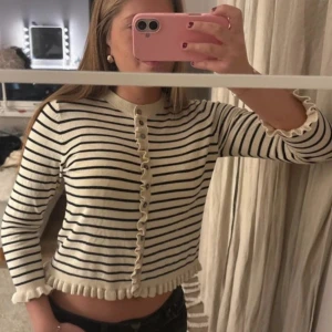 Randig kofta med volangkant - Supersöt kofta i vit och svart med smala ränder och volangdetaljer vid ärmslut och nederkant. Tröjan har lång ärm, knappar framtill och är croppad i modellen. Perfekt för dig som gillar en gullig och trendig stil.