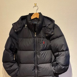 Svart pufferjacka från Polo Ralph Lauren - Svart pufferjacka från Polo Ralph Lauren med röd logga på bröstet. Jackan har huva, snörning och tvåvägsdragkedja med RL-detaljer. Fylld med dun och har en clean, boxig siluett. Perfekt för kalla dagar och har praktiska fickor framtill.