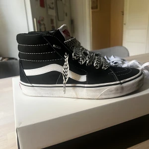 Svarta Vans Sk8-Hi sneakers - Svarta Vans Sk8-Hi sneakers med vit sidorand och kontrastsydda detaljer. Skorna har högt skaft, platt sula och snörning med svartvita zigzag-mönstrade skosnören samt extra vita skosnören. Materialet är en mix av mocka och canvas för en cool streetstil. Säljer då dom va för små på mig! Snöret har jag själv lagt till men orginalsnöret kommer också med! Använda 2 gånger💕