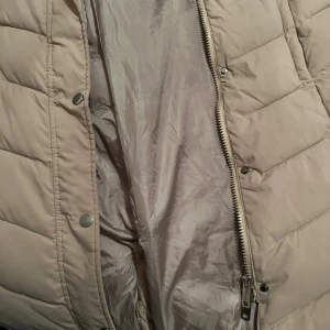 Beige lång pufferjacka White Icy XL - Säljer en beige lång pufferjacka från White Icy i storlek XL. Jackan har huva med svart fuskpäls, dragkedja och tryckknappar framtill, samt två sidofickor med dragkedja. Insidan är fodrad med mjuk pälsimitation för extra värme. Perfekt för kalla dagar.