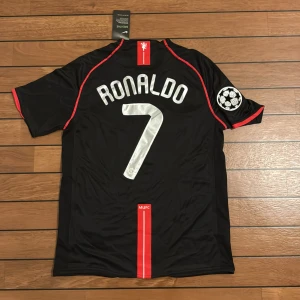 Manchester United Ronaldo 7 svart tröja - Säljer en svart Manchester United fotbollströja med Ronaldo 7 på ryggen. Tröjan har röda detaljer, Champions League-märke på ärmen och AIG som sponsor på bröstet. Tillverkad i lätt och ventilerande Nike FitDry-material.