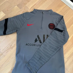 Grå långärmad PSG träningströja Nike - Snygg grå långärmad träningströja från PSG med svart och rött tryck. Tröjan har halv dragkedja, diskret mönster på ärmarna och svart Nike-logga samt PSG-märke på bröstet. Perfekt för fotbollsträning eller chill.
