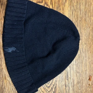 Marin blå stickad mössa från Polo Ralph Lauren - Marin blå stickad mössa från Polo Ralph Lauren med ribbad kant och diskret broderad logga på sidan. Klassisk och enkel design som passar perfekt när det är kallt ute. Mjuk och skön kvalitet i ullblandning.
