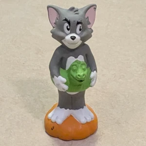 Tom figur av mjuk plast , Tom & Jerry, 1993 - Figur av Tom från Tom & Jerry, stående på orange bas med grön badring. Färgerna är klara, mindre färgslitage syns på öronen. Perfekt för samlare eller användas som leksak eller som dekoration. Bra skick.  Ett mörkt kort sträck finns på det orangea som Tom står på.