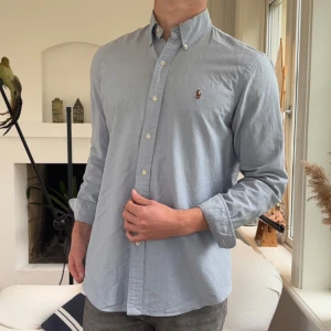 Ljusblå skjorta från Ralph Lauren - Klassisk ljusblå skjorta från Ralph Lauren med button-down krage och broderad logga på bröstet. Skjortan har lång ärm, custom fit och stängs med vita knappar. Perfekt för en clean och stilren look.
